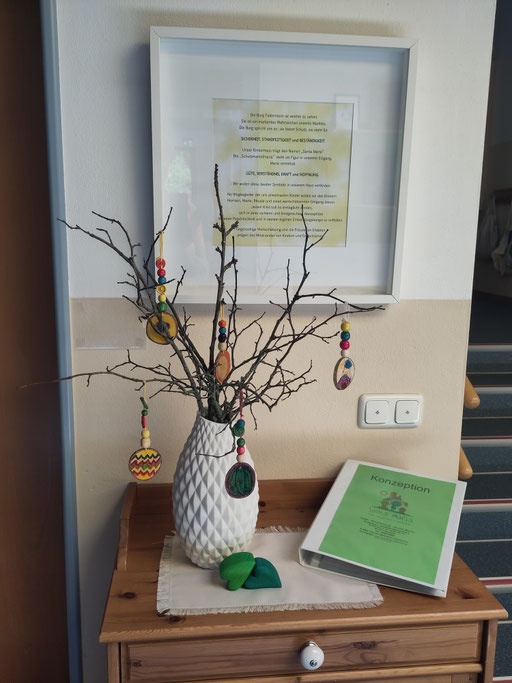 Ein gebundener Ordner mit der Aufschrift „Konzeption“ liegt auf einem Tisch neben einer Vase mit Zweigen und buntem Schmuck.